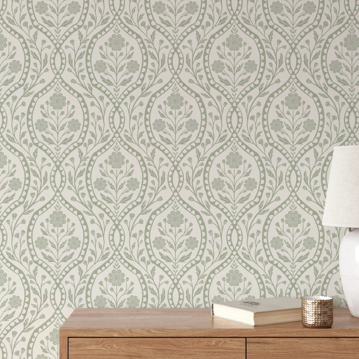 a wooden dresser with a lamp and a book on top of it, set against a wall with a repeating pattern of green floral designs.