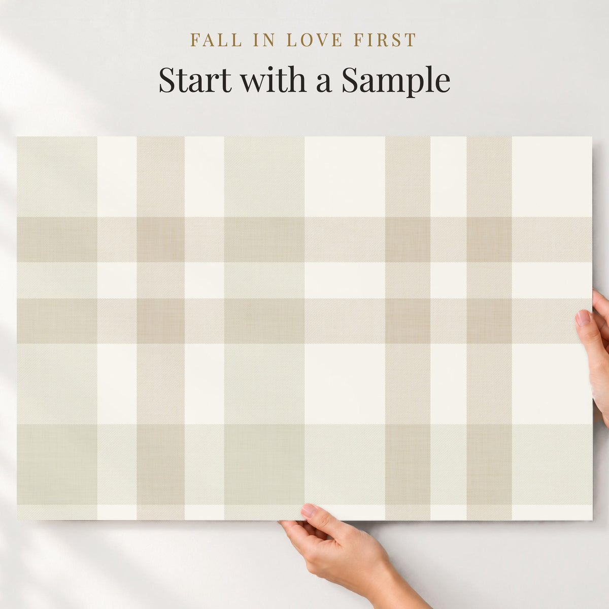 Hands holding a large sample of beige and cream Hollis plaid wallpaper against a wall