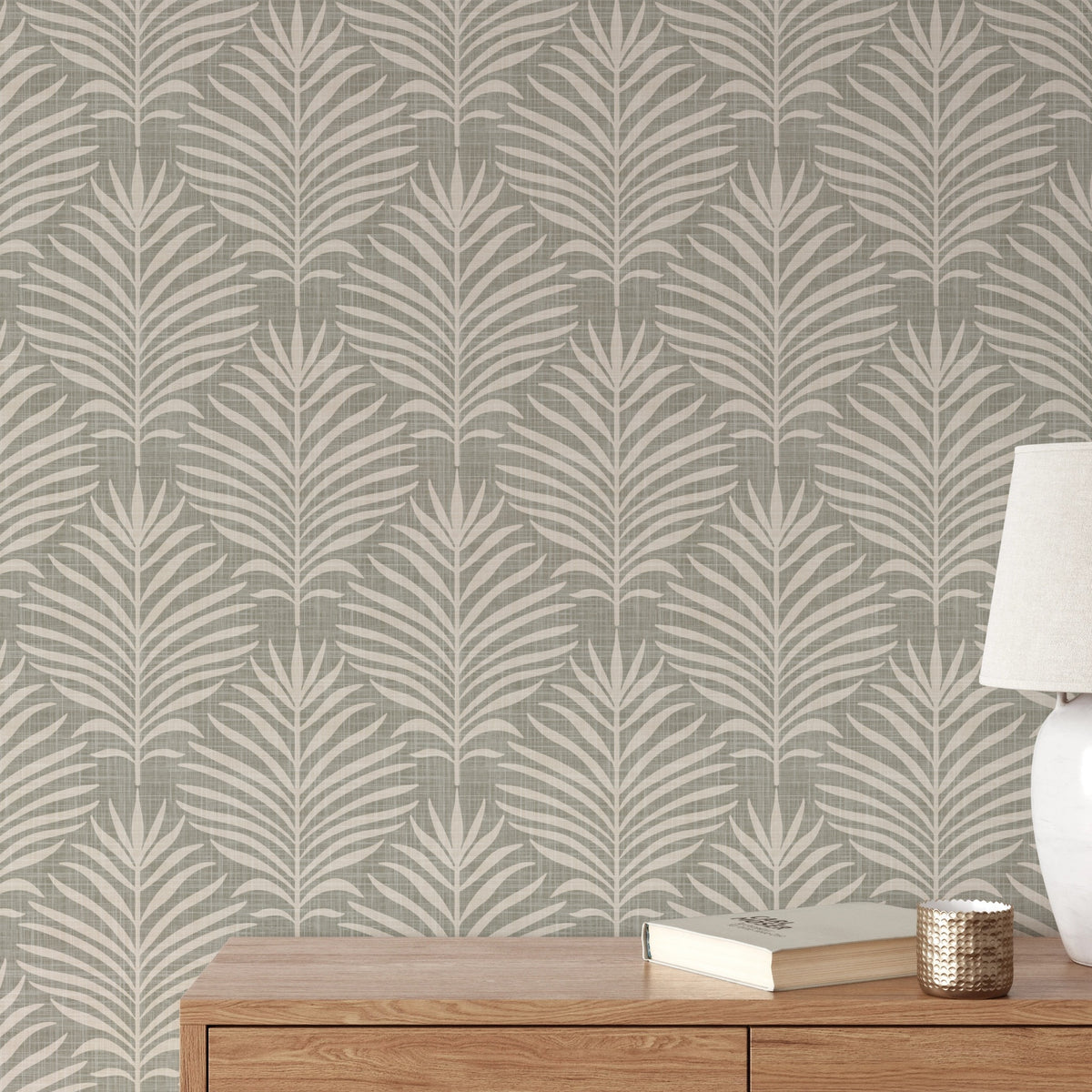 a wooden dresser with a lamp and a book on top, set against a wall with a repeating pattern of palm leaves in a neutral color palette.