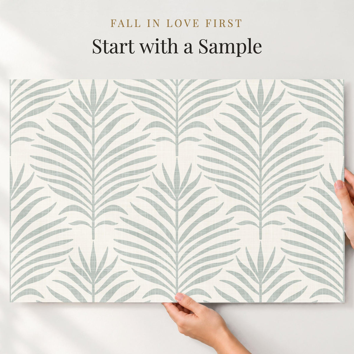 Hands holding a large sample of gray green and white Palm Breeze palm leaf wallpaper against a wall