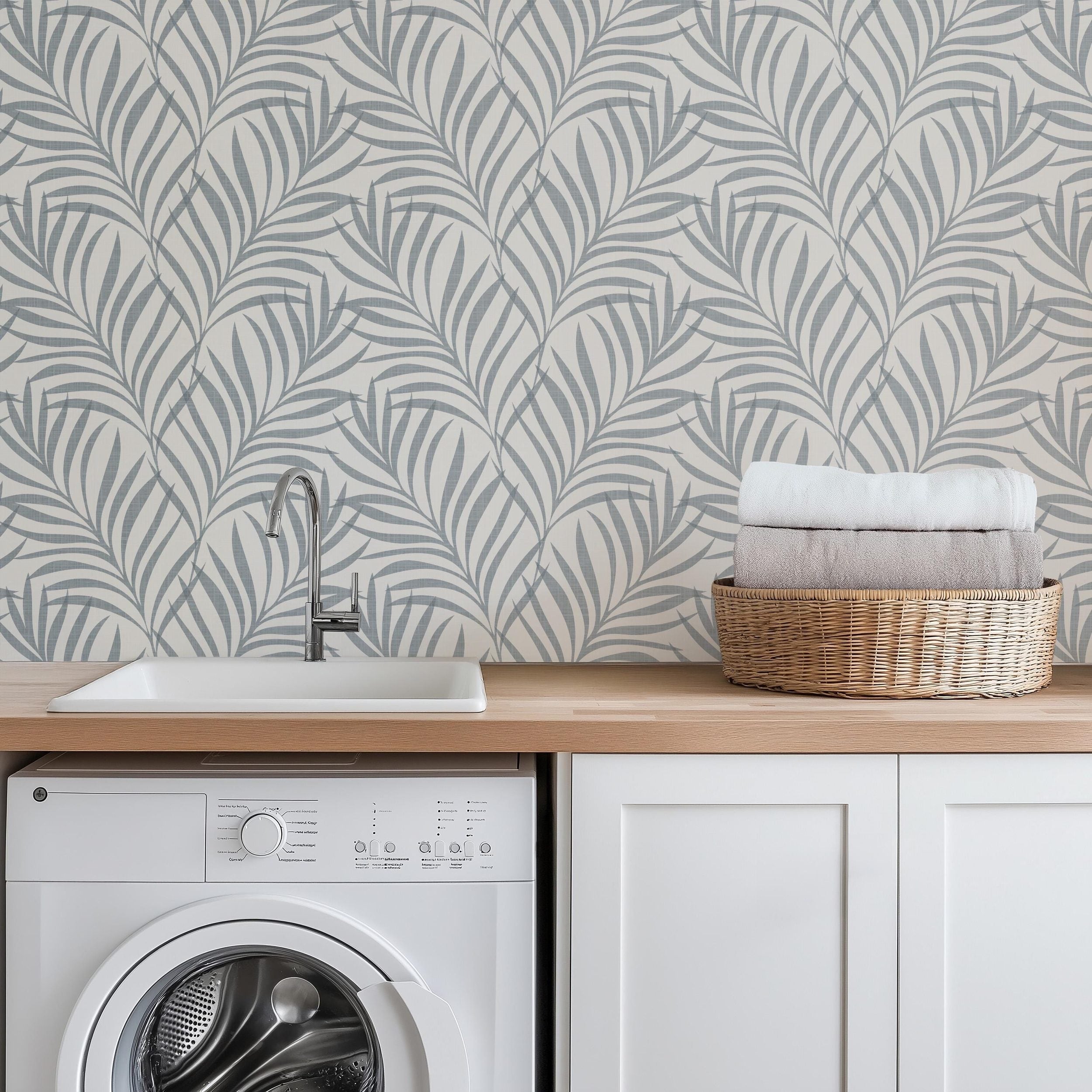 blue and off white palm leaf removable wallpaper available in peel and stick with a faux linen texture for japandi, coastal, and boho decor styles. featured as laundry room wallpaper behind washer.